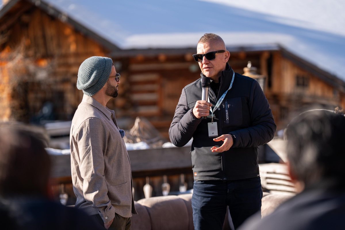 Oliver Camponovo at the microphone, in conversation with a second speaker before an audience, outside a timber chalet in the snow